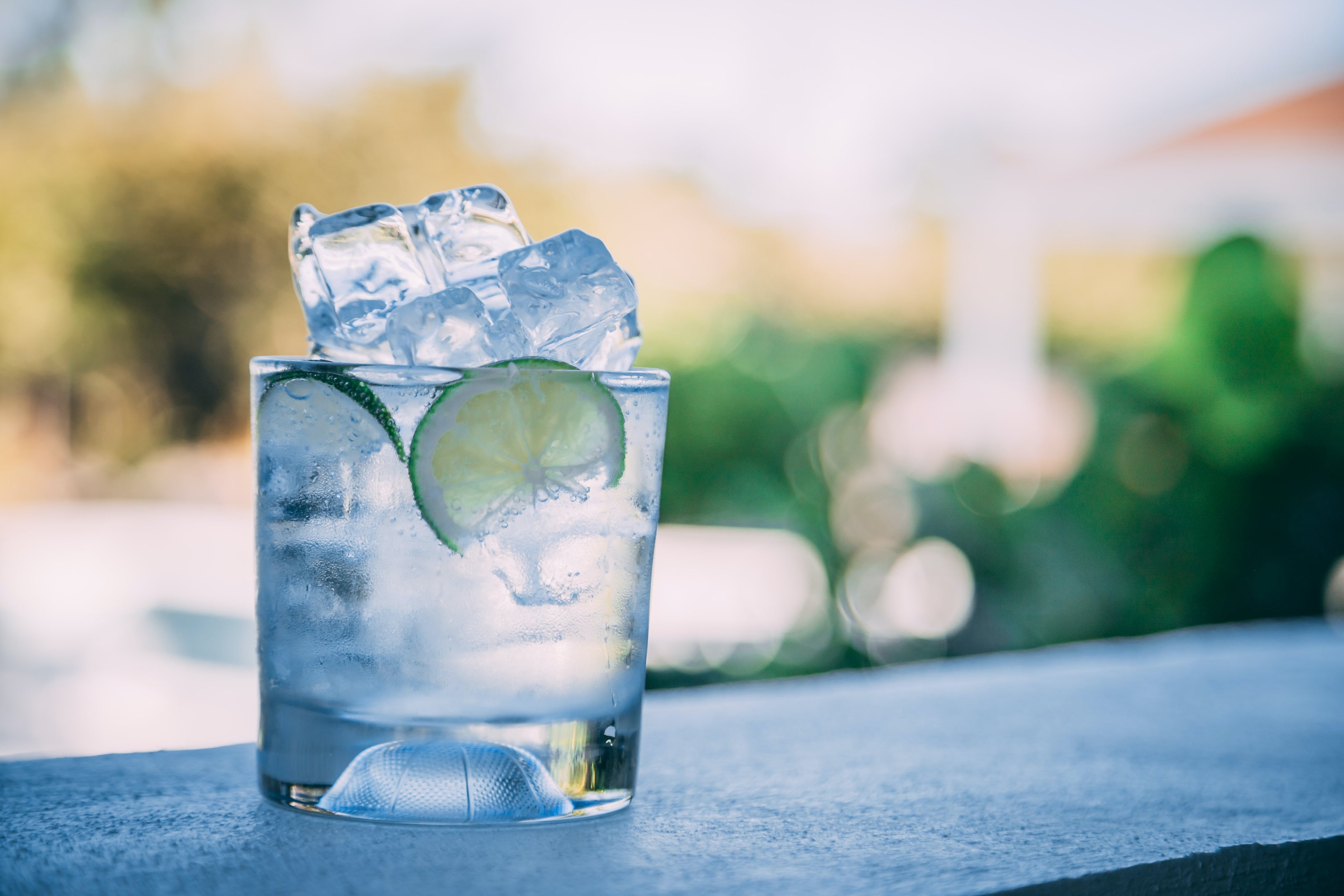 Clear glass with a blue drink, lime slices, and ice cubes on a blurred outdoor background